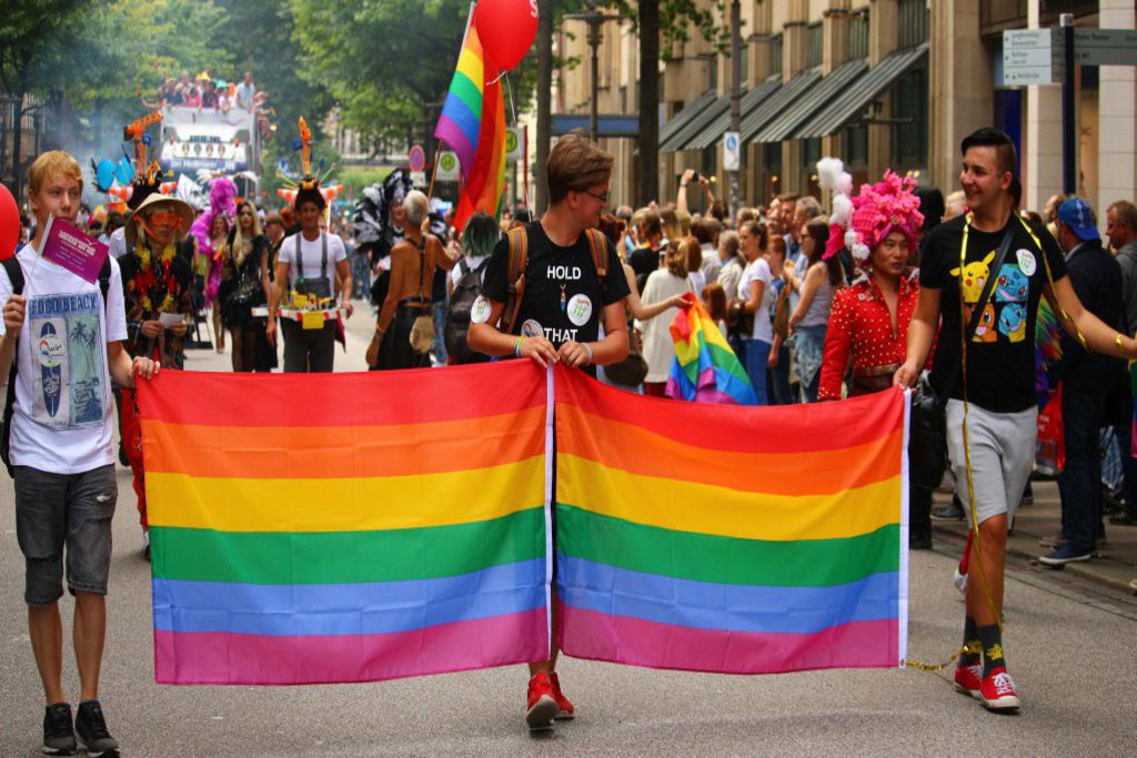 Drei Männer halten zwei Regenbogenflaggen und marschieren mit einer Menschenmenge.