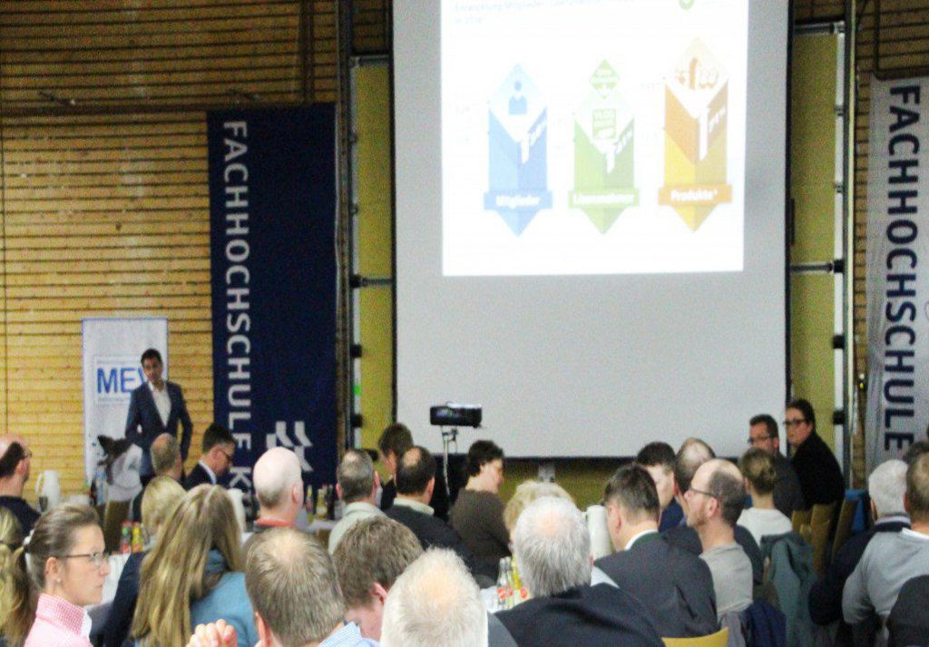 Ein Publikum, dass an vertikal zur Bühne ausgerichteten Tischen sitzt wartet auf den Beginn des Vortrags.