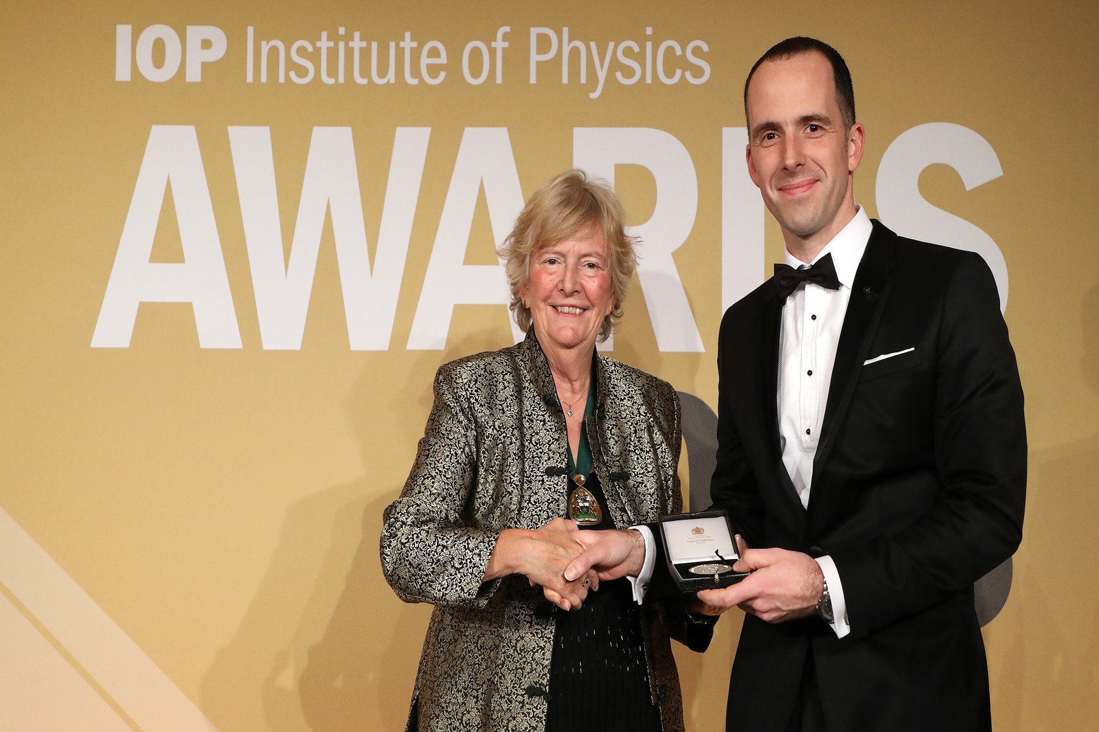 eine frau und ein Mann reichen sich die Hände. Der Mann hält eine Auszeichung in der Hand.