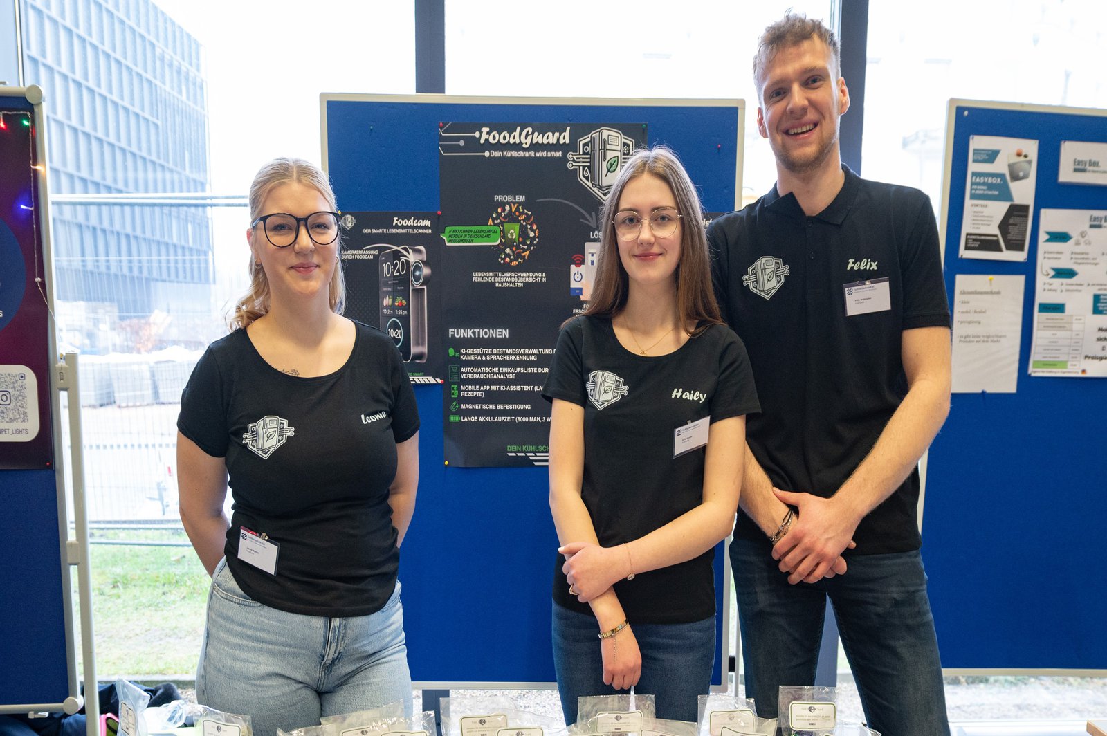 zwei Schülerinnen und ein Schüler des RBZ Steinburg stehen beim Finale der StartUp Challenge 2025 an ihrem Tisch im Audimax der FH Kiel