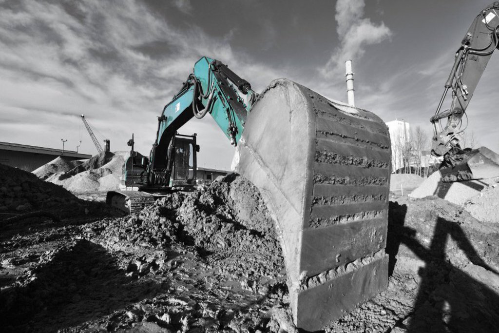 Eine Schwarz-Weiss-Aufnahme auf der lediglich ein Bagger in grün gefärbt ist. Die Baggerschaufel lädt gerade Sand ab.