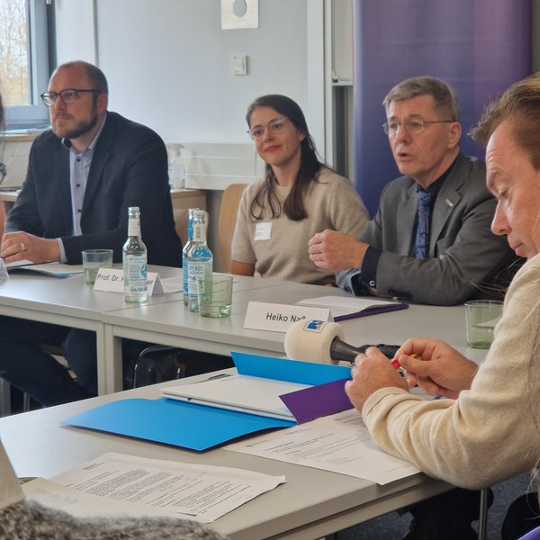 Aufnahme einer Pressekonferenz, an einem Tisch an der Stirnseite sitzen zwei Männer und eine Frau. Seitlich davon ein Reporter mit einem Mikrofon
