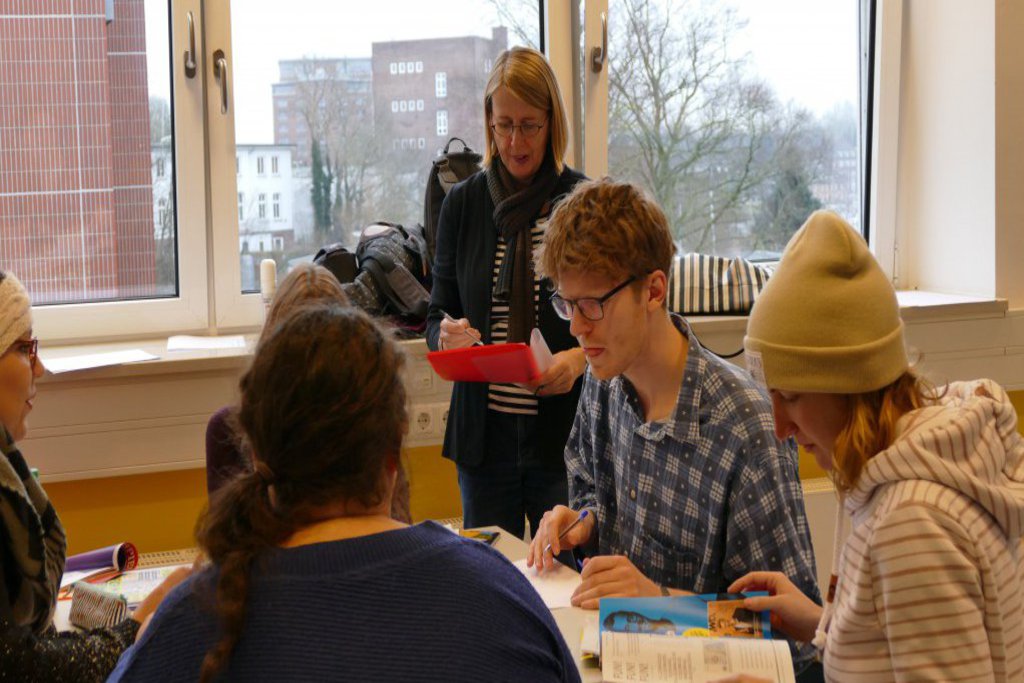 Fünf Studierende sitzen an einem Tisch. Ein Lehrkörper steht daneben und macht Notizen.