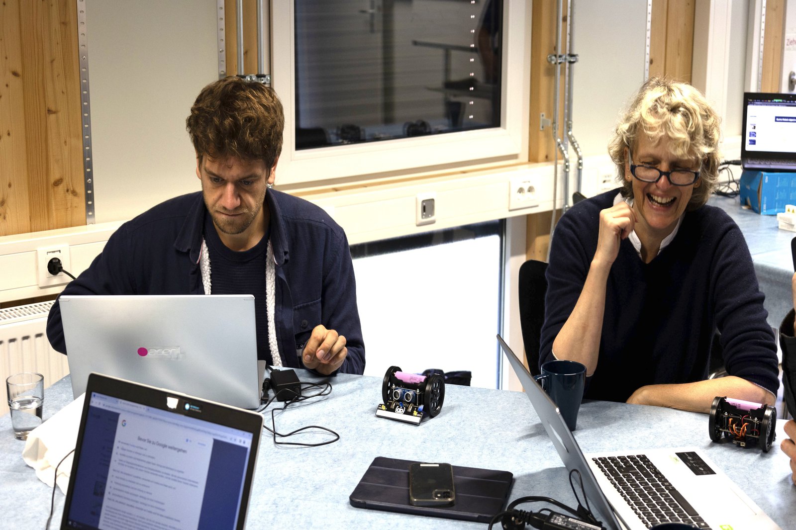 Ein Mann und eine Frau sitzen gemeinsam an einem Tisch. Auf dem Tisch stehen Laptops und ein kleiner Roboter.