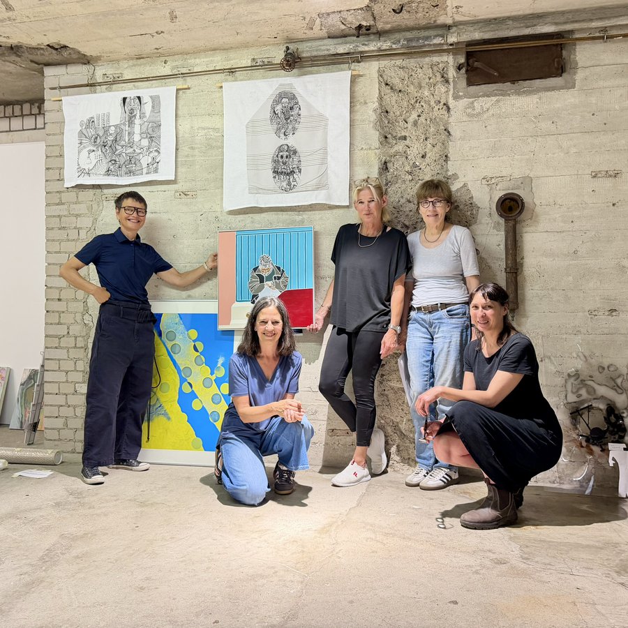 5 Frauen gruppieren sich um ein paar Kunstwerke, die an einer rauen Betonwand hängen