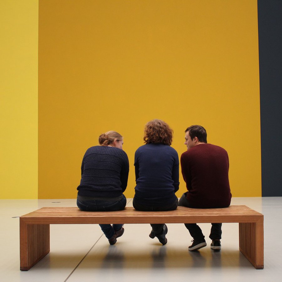 Drei Menschen sitzen auf einer Bank im Museum. Sie schauen auf eine gelbe Wand.