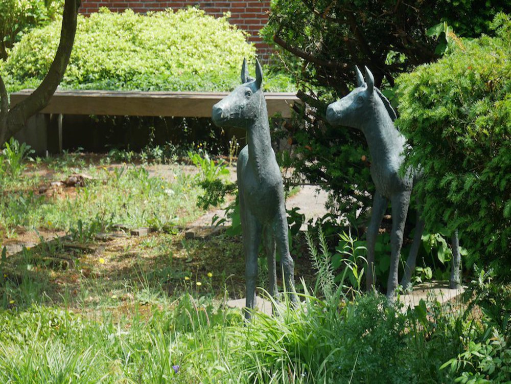 Zwei Pferdeskulpturen stehen auf einer Grünfläche, in der Nähe eines Buschs.