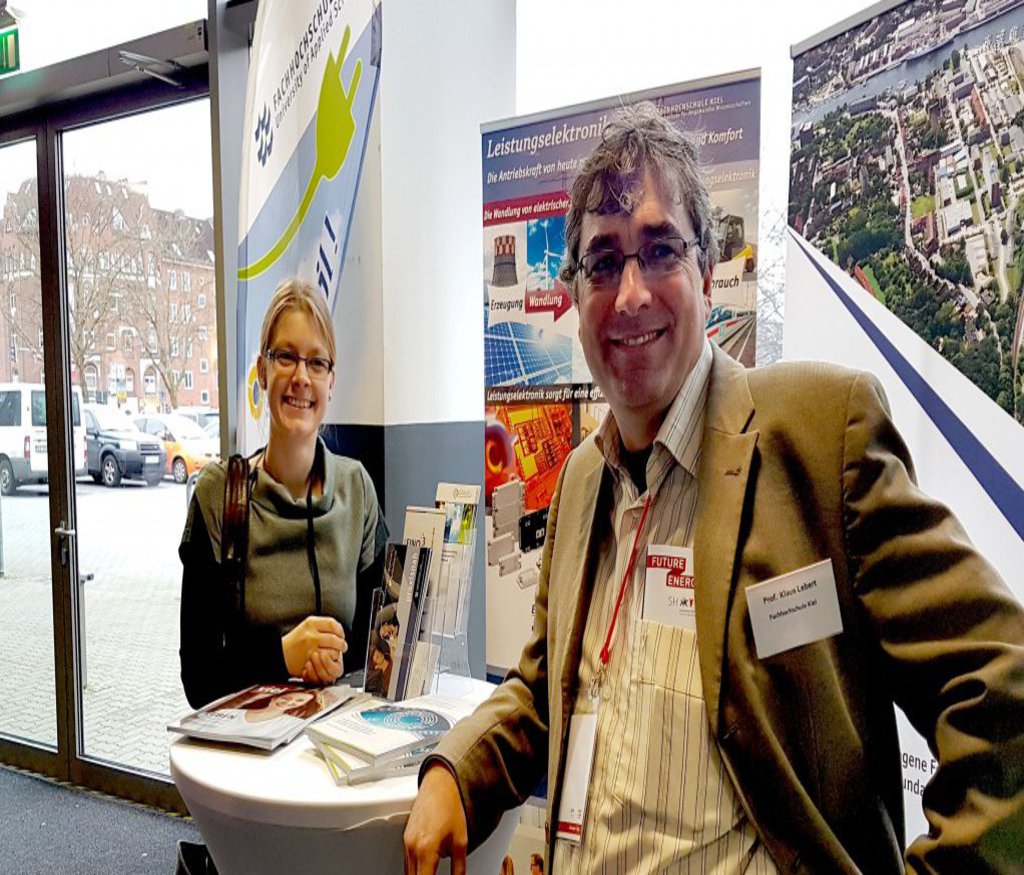 Ein Mann und eine Frau stehen an einem Infotisch und lächeln freundlich in die Kamera.