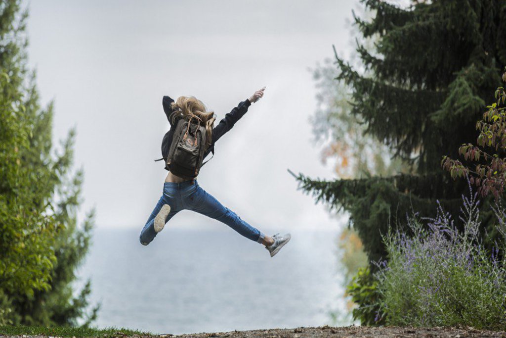 Eine Frau springt, auf dem Weg ans Meer, freudig mit beiden Beinen in die Luft.