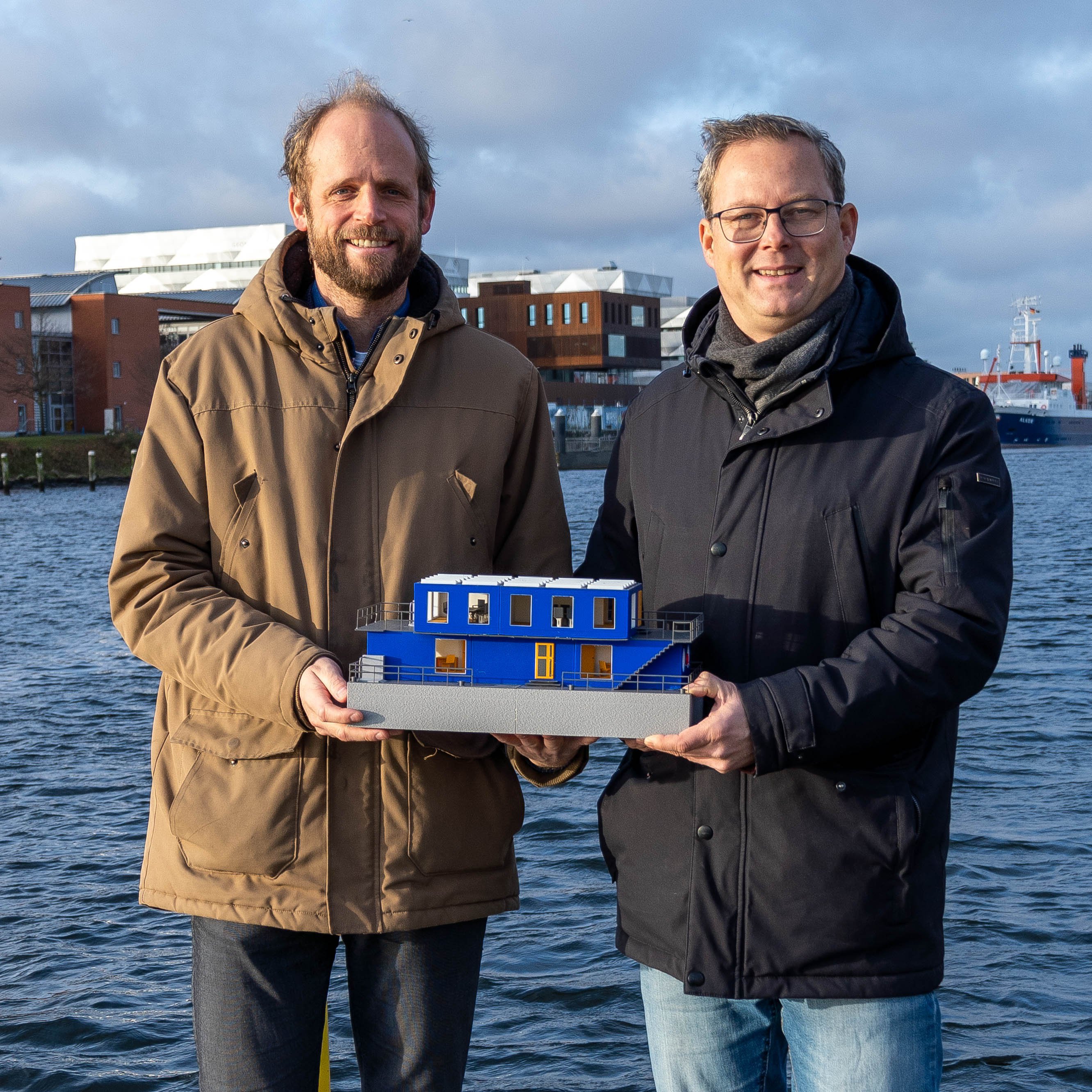zwei Männer stehen nebeneinenader und halten das Modell eines blauben rechteckigen Hausboots. Im Hintergrund ist Wasser zu sehen und das gegenüberliegende Uferist  