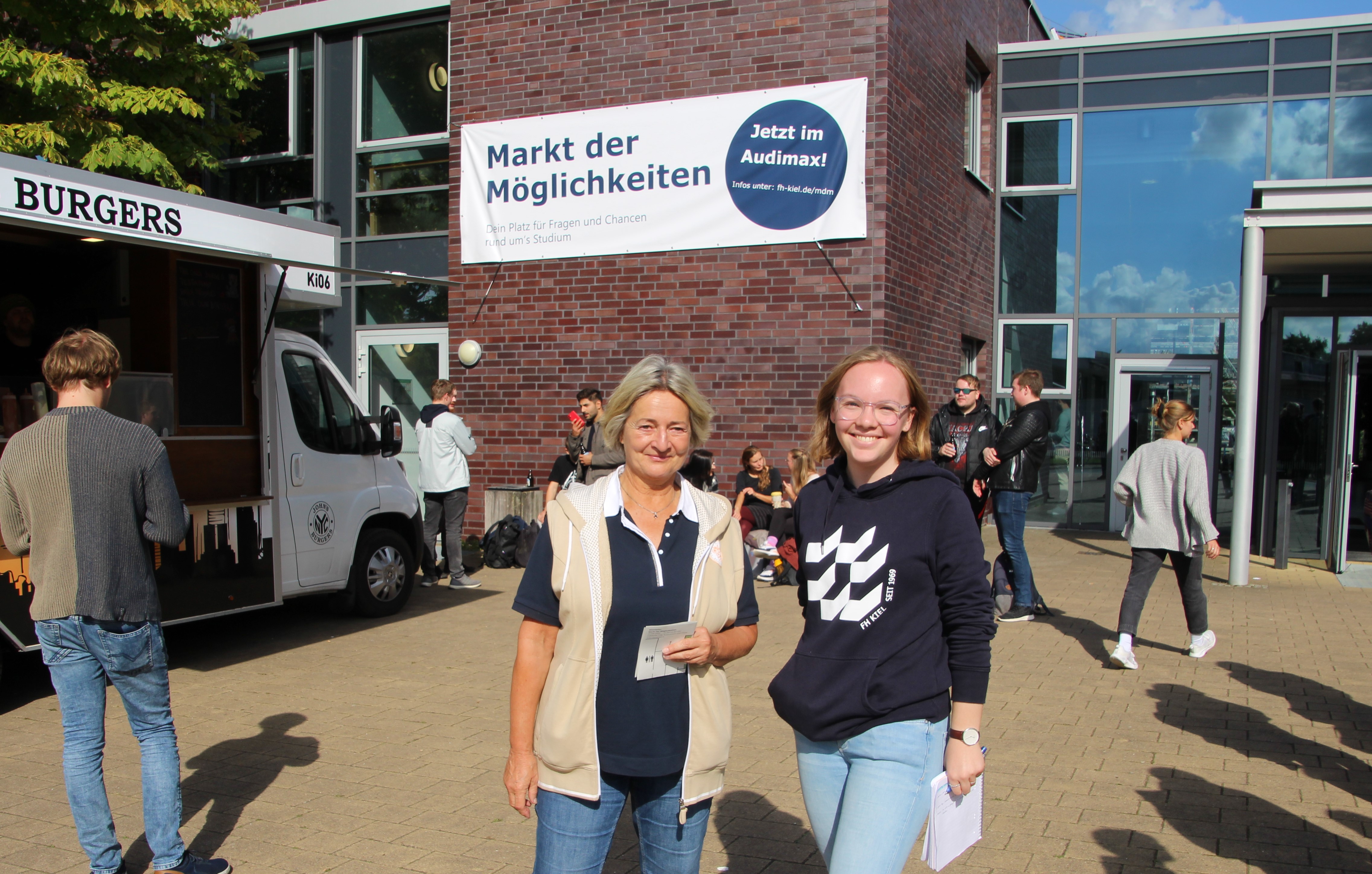 Zwei Frauen schauen lächelnd in die Kamera, hinter ihnen hängt ein Banner an einem Gebäude. Aufschrift: Markt der Möglichkeiten