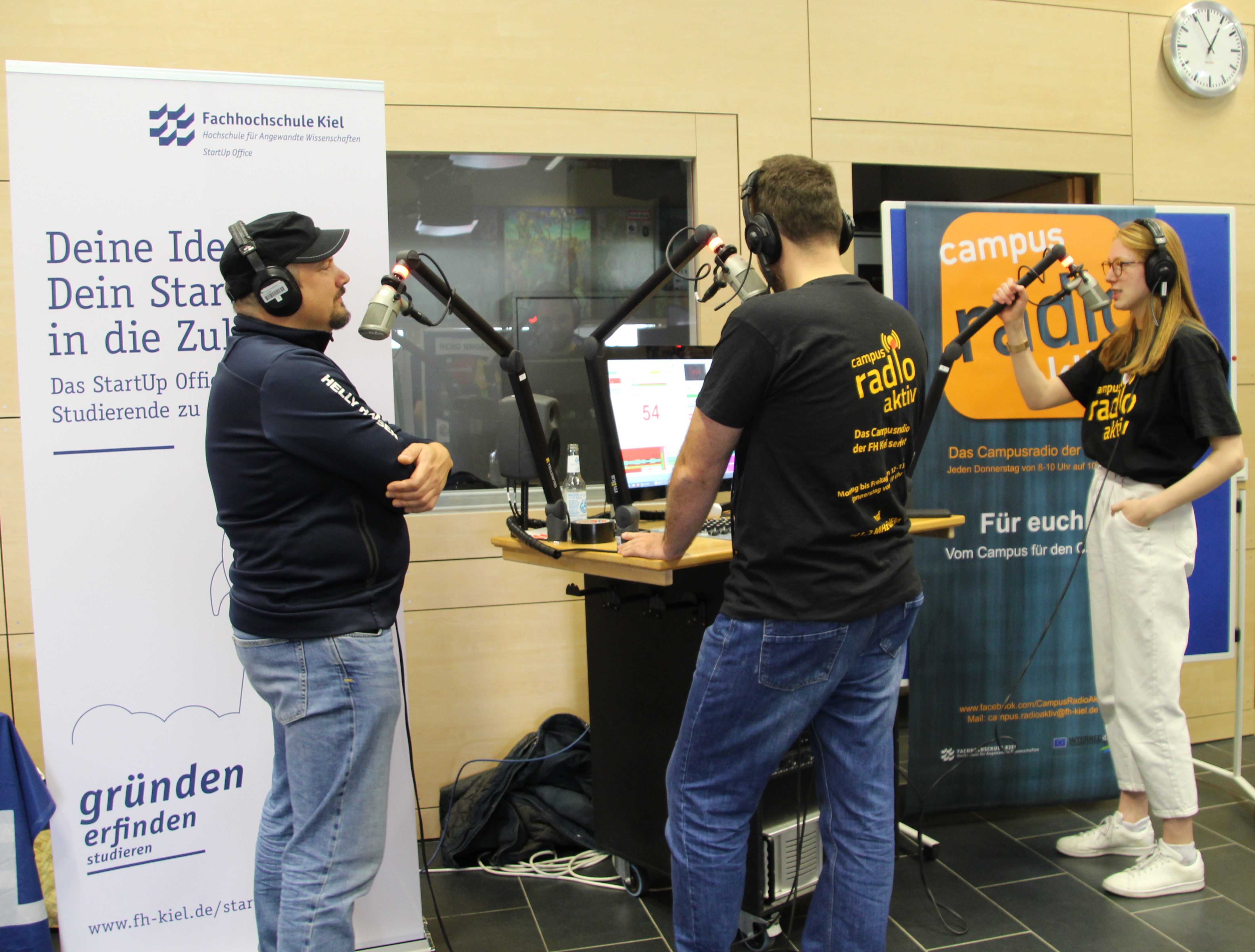 Zwei Männer und eine Frau stehen vor Radiomikrofonen, sie tragen Kopfhörer