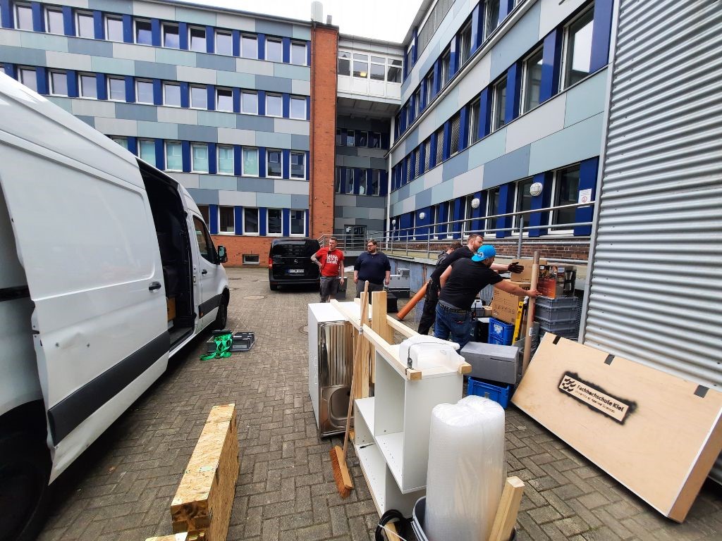 Foto: Eisele ein Transporter, daneben Menschen, die einladen; dahinter ein Gebäude