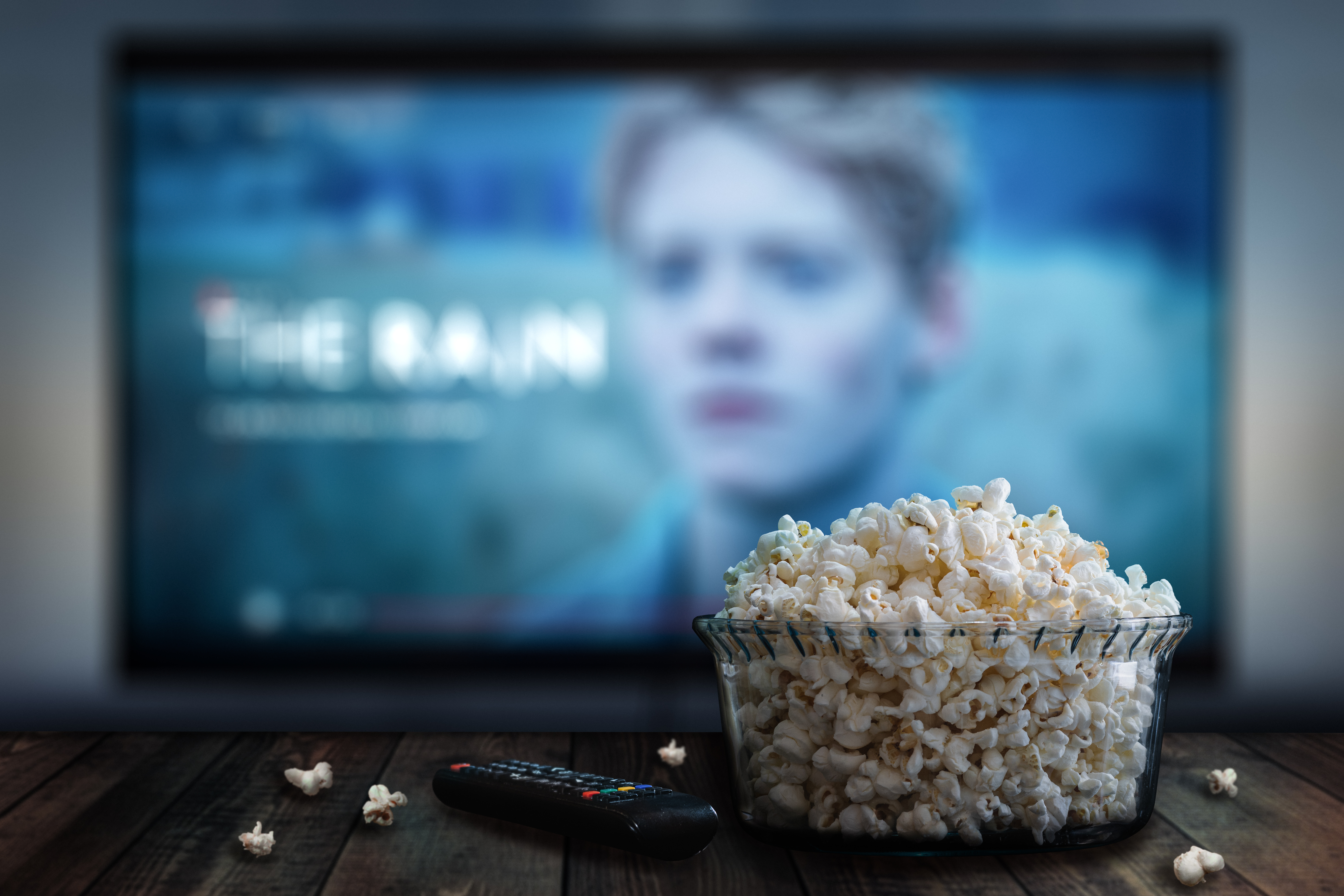 Video streaming app on tv screen behind a bowl of popcorn and a eine Schale mit Popcorn steht auf einem Tisch neben einer Fernbedienung, dahinter verschwommen ein TV-Bildschirm