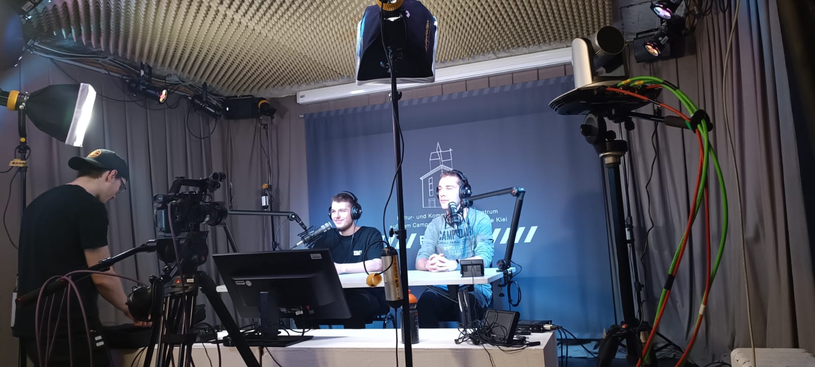 auf einer Bühne hinter einem Tisch sitzen zwei Männer. Sie werden von einem Scheinwerfer im bläulichen Licht angestrahlt und eine Kamera ist auf sie gerichtet.