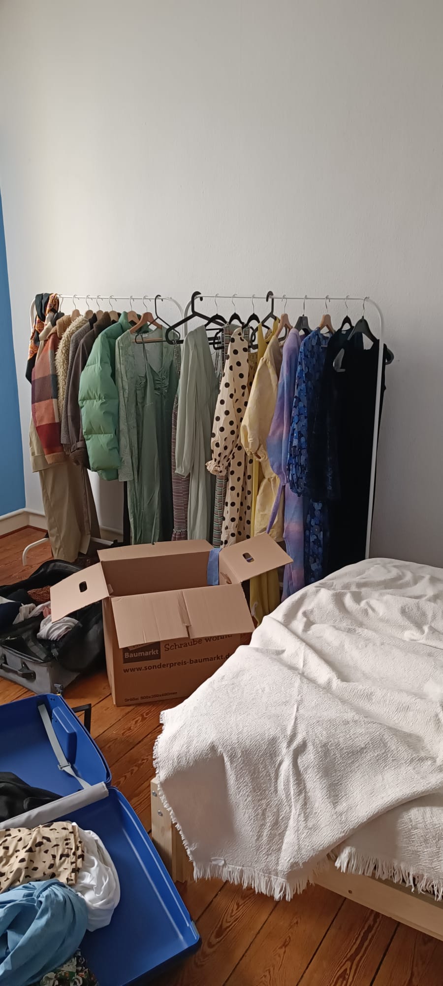 Ein leeres Zimmer mit blauen und weißen Wänden. Man sieht einen Umzugskarton, einen Koffer und eine Kleiderstange.