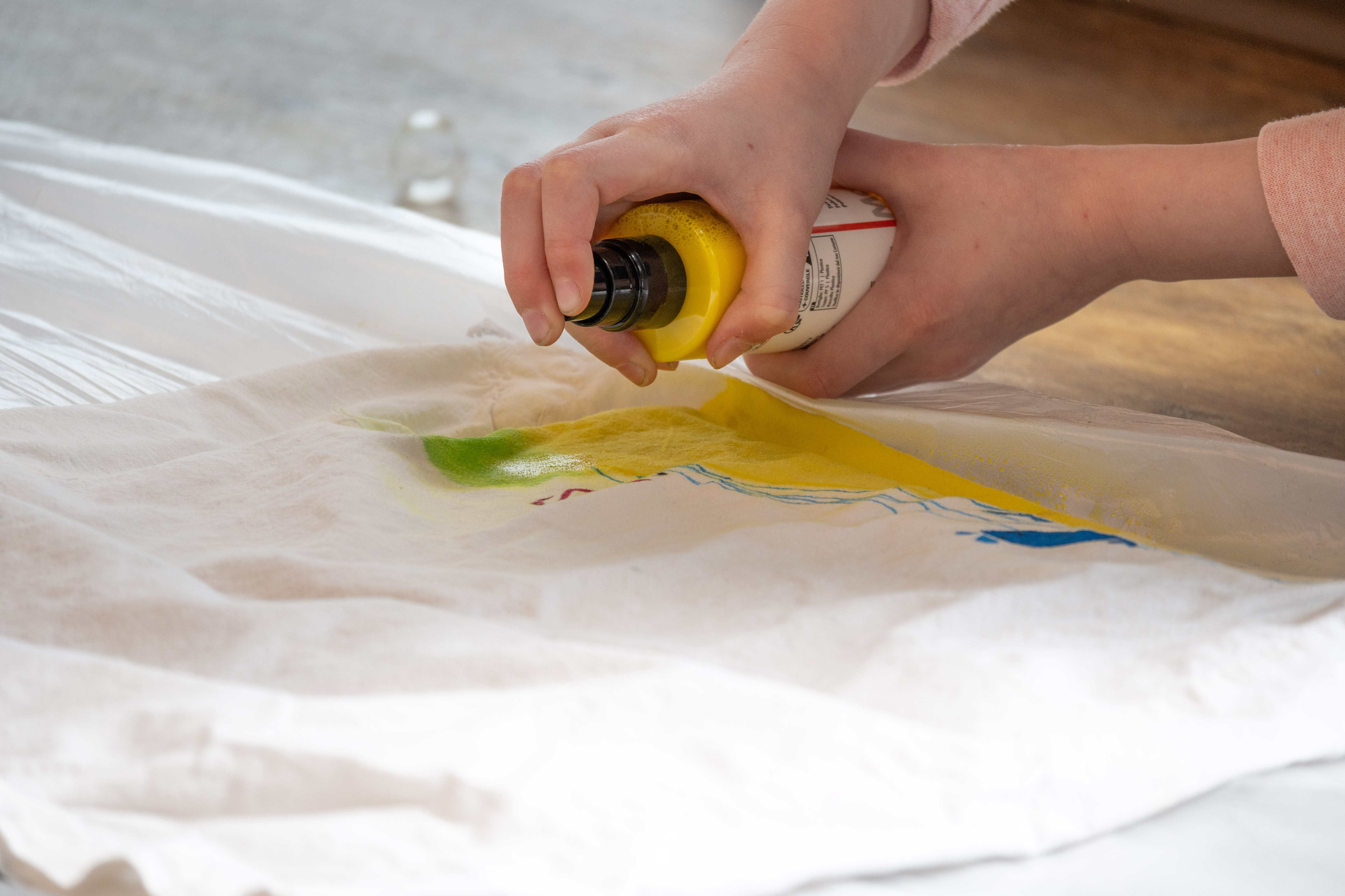 zwei Hände halten eine Sprühflasche und besprühen ein T-Shirt mit Farbe.