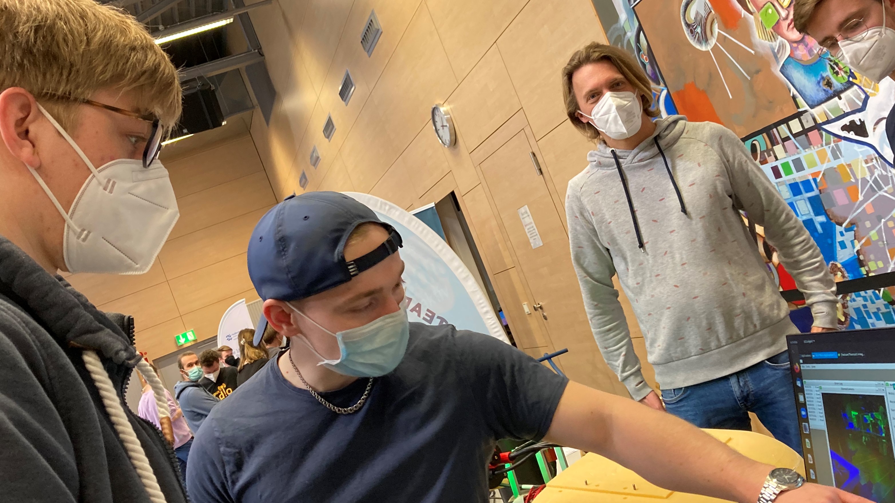 Ein junger Mann schaut auf einen Bildschirm, auf dem ein anderer junger Mann etwas zeigt. Im Hintergrund sind zwei weitere junge Männer zu sehen.