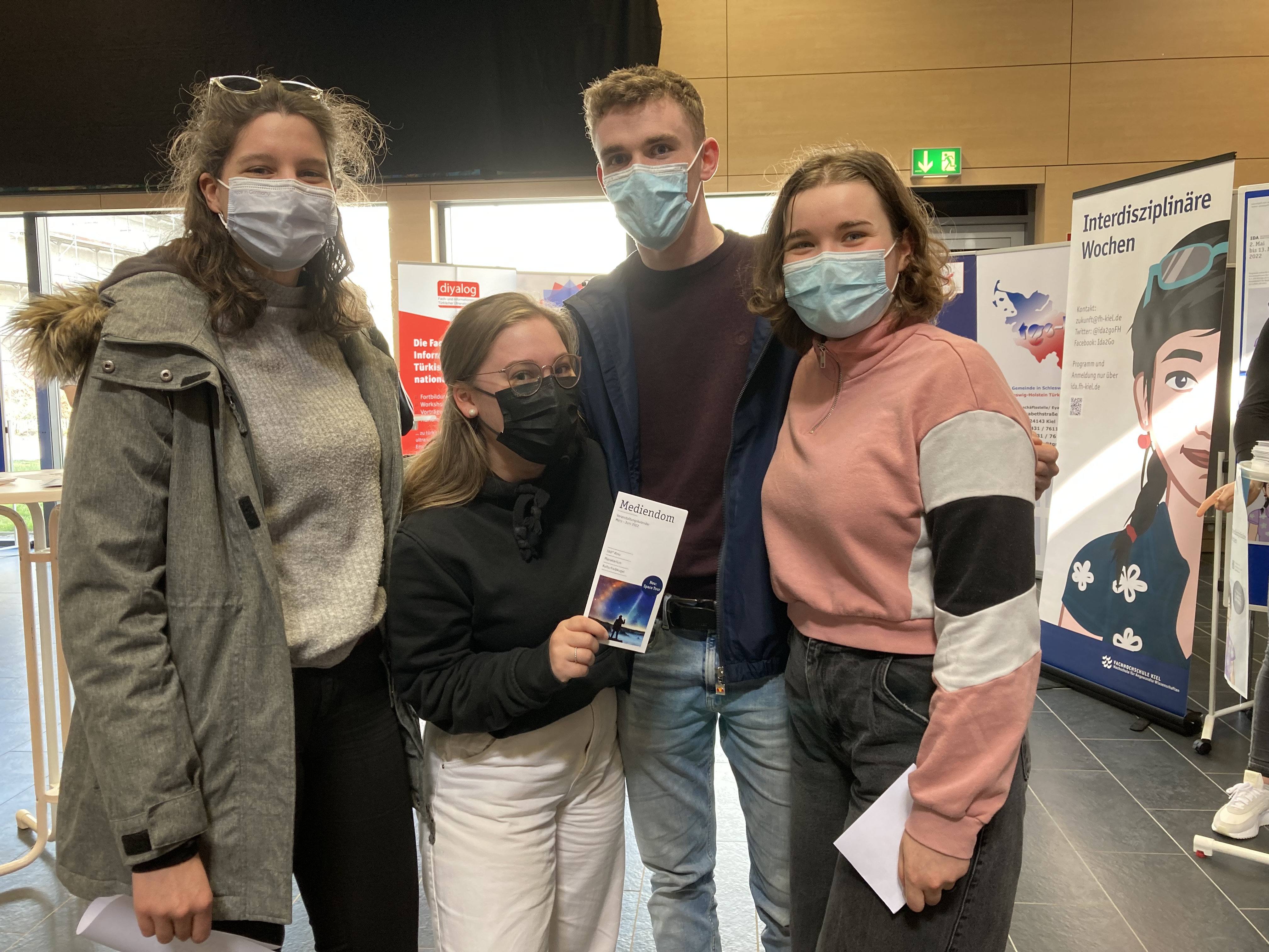 Drei junge Frauen und ein junger Mann stehen dicht nebeneinander. Eine der Frauen hält eine Infobroschüre in die Kamera.