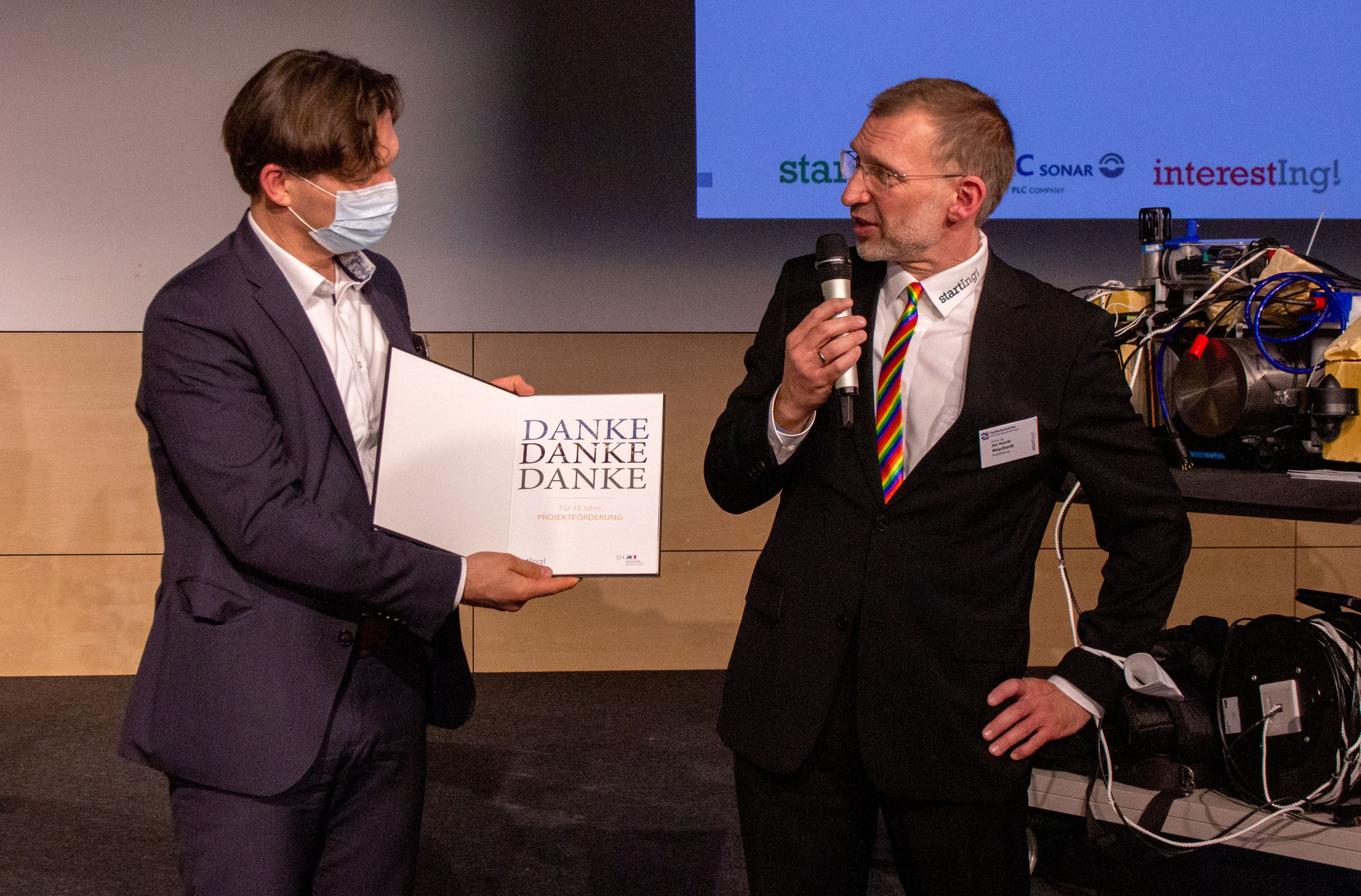 Zwei Männer auf der Bühne, einander zugewandt: Der Mann links im Bild hält eine Danksagungskarte, der Mann rechts im Bild hält ein Mikrofon.