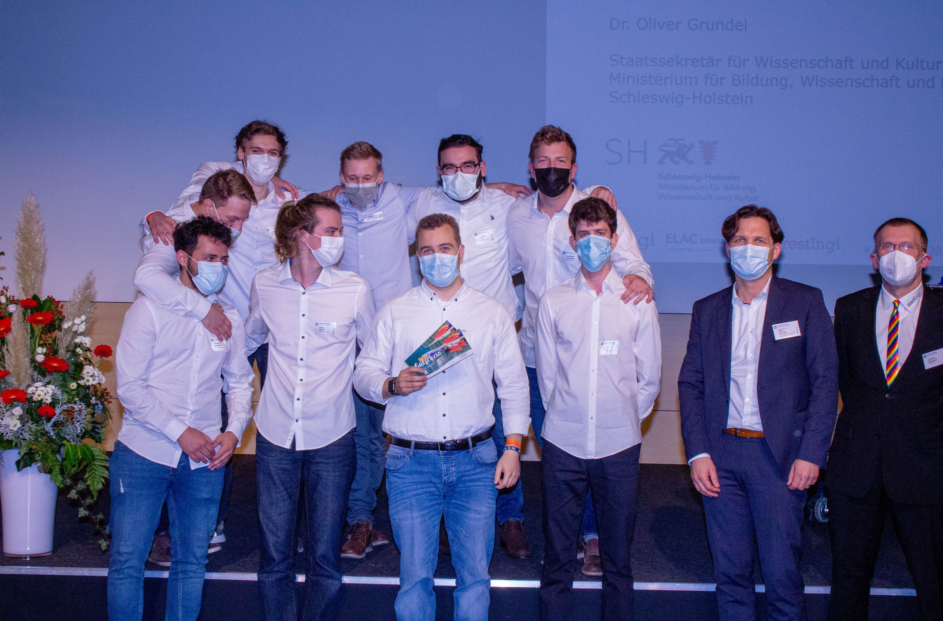 Eine Gruppe junger Männer in weißen Hemden auf einer Bühne, zwei Herren in Anzug daneben