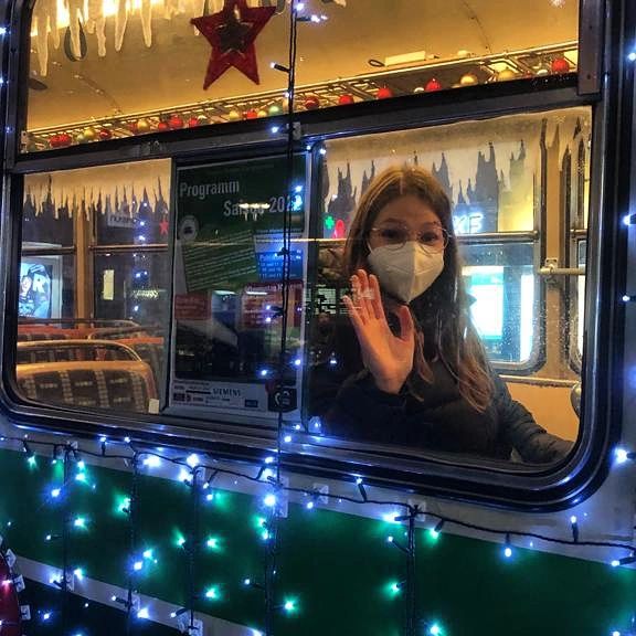 Jana sitzt mit Maske in einer Straßenbahn und winkt. Die Straßenbahn ist geschmückt mit Lichterketten