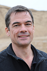 Ein Mann mit schwarzem T-Shirt steht auf einer Baustelle und lächelt in die Kamera.