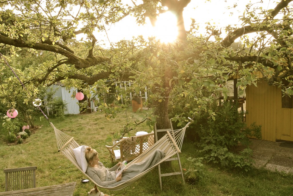 Eine Frau liegt in einem hellen grünen Garten, in einer Hängematte und liest ein Buch.