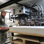 Durch die Druckerpresse hindurch ist ein Mann bei der Arbeit zu sehen.