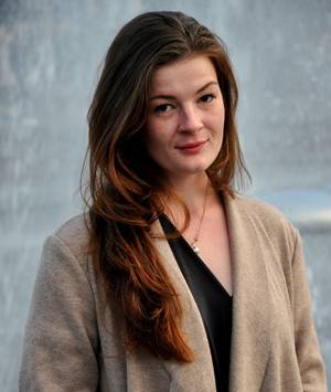 Eine Frau in brauner Jacke neigt den Kopf zur Seite und schaut in die Kamera.