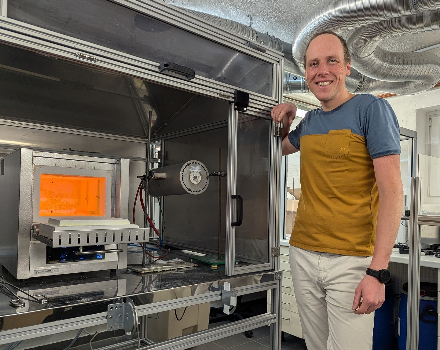 Der Mann in der goldgelben Arbeitskleidung steht lächelnd neben einem industriellen 3D-Drucker in einem Techniklabor.