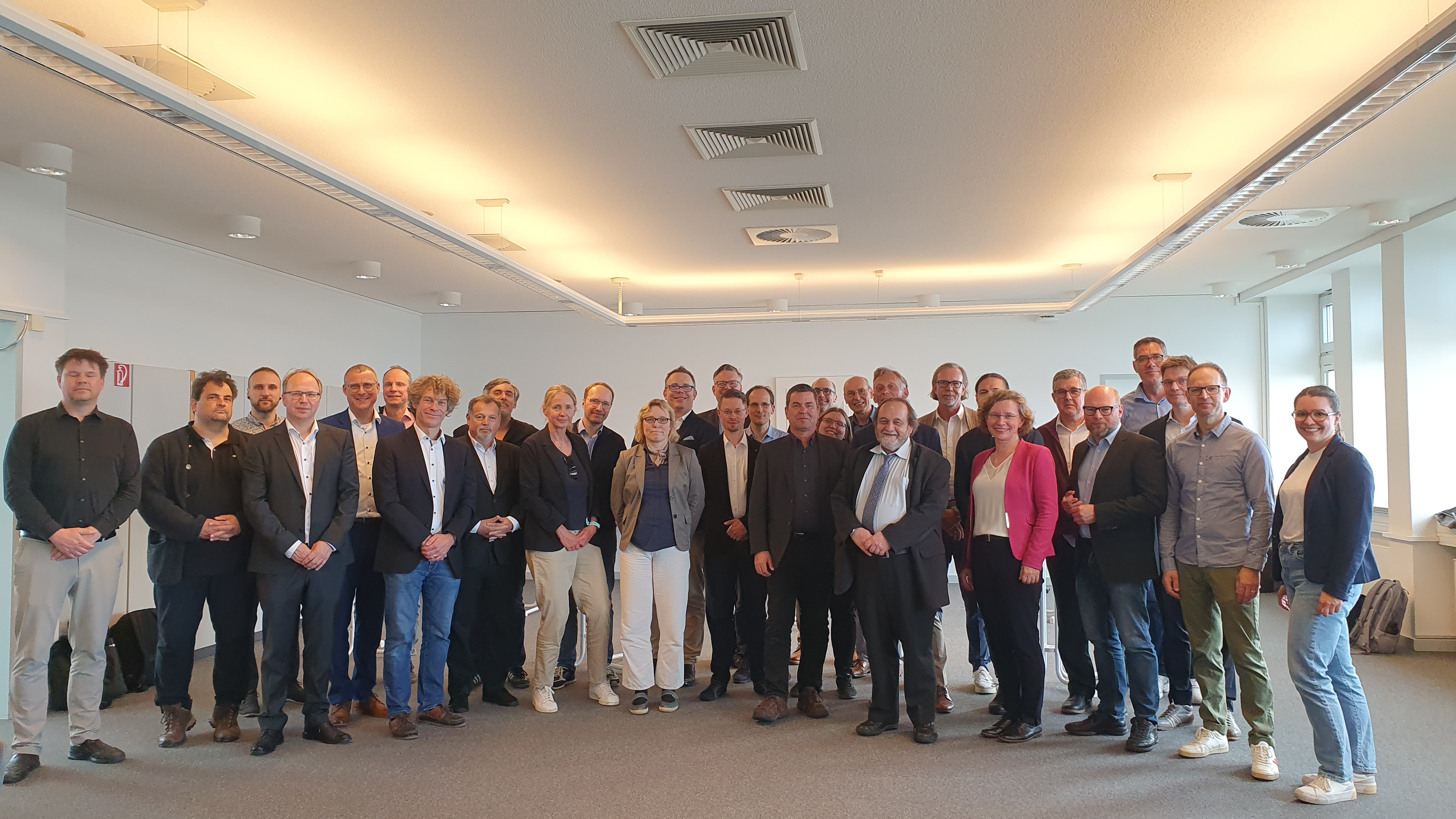 Auf dem Bild sind 31 Personen zu sehen, die als Gruppe im Halbkreis stehen. Sie sind business casual gekleidet und lächeln in die Kamera.