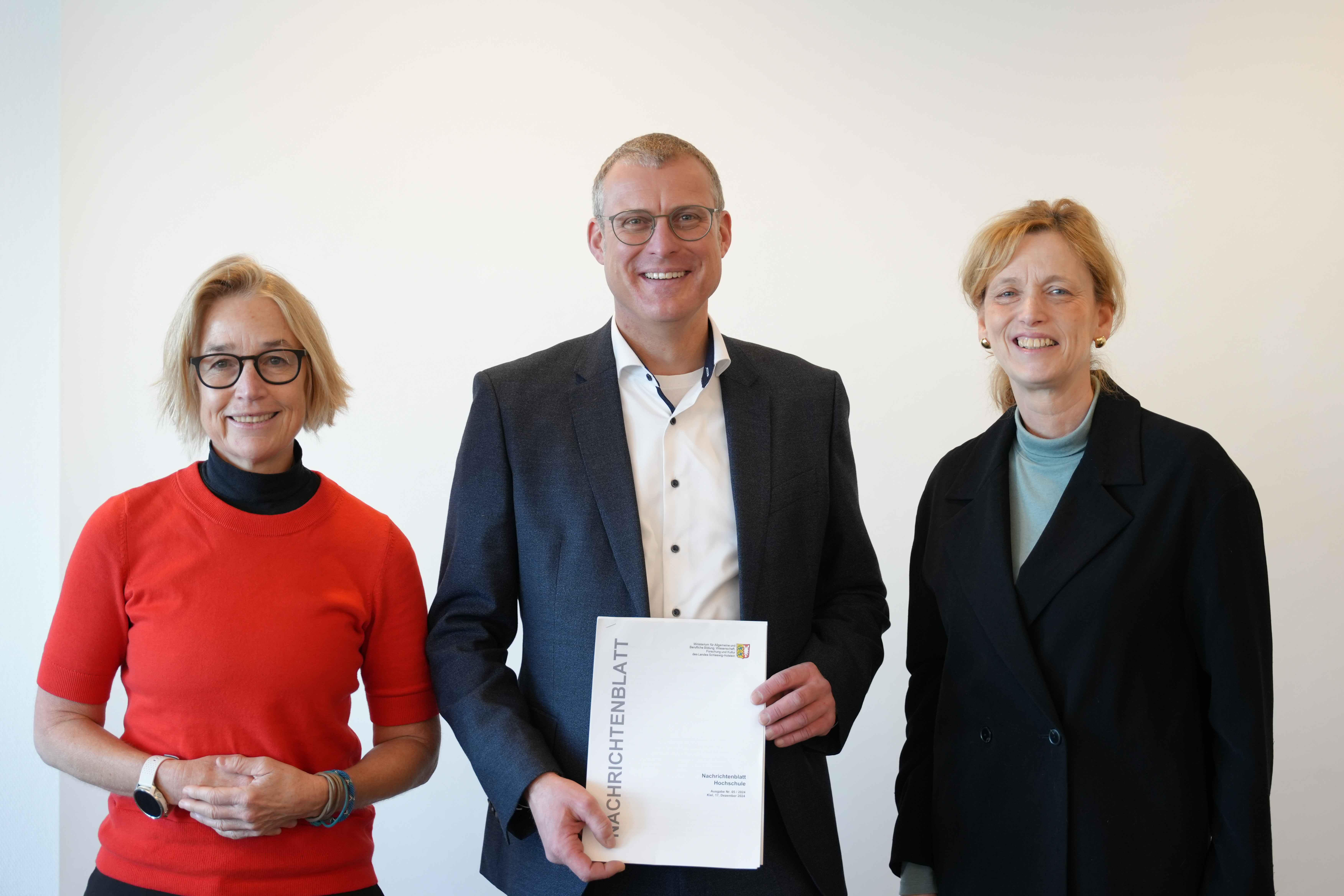 Auf dem Bild sind von links nach rechts zu sehen: Prof. Dr. Christiane Hipp, Prof. Dr. Björn Christensen, Ministerin Karin Prien. Alle drei lächeln in die Kamera. Björn Christensen hält das Nachrichtenblatt Hochschule in der Hand.